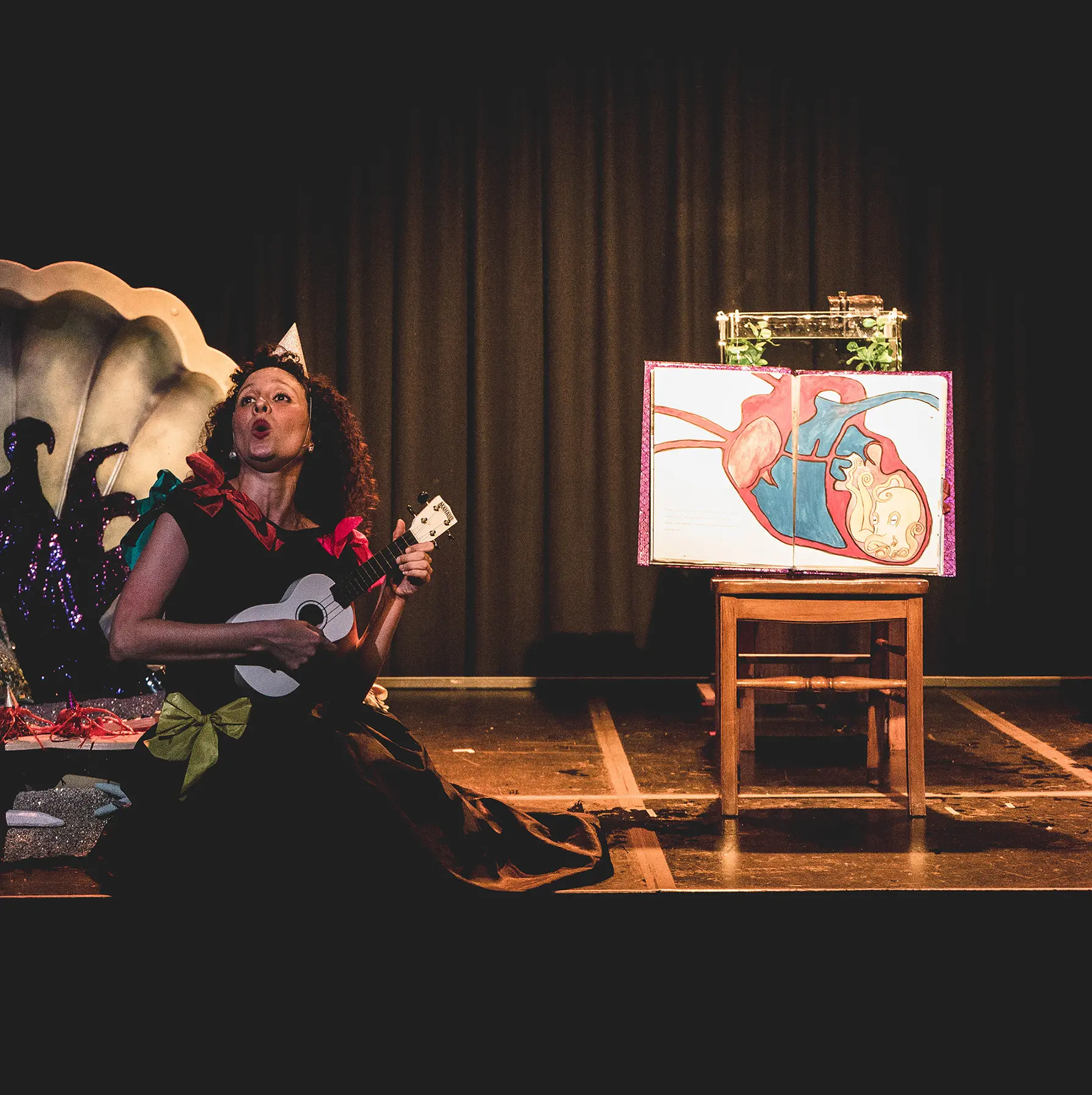 A woman sings and plays the ukulele beside a large picture book showing an octopus trapped in a human heart