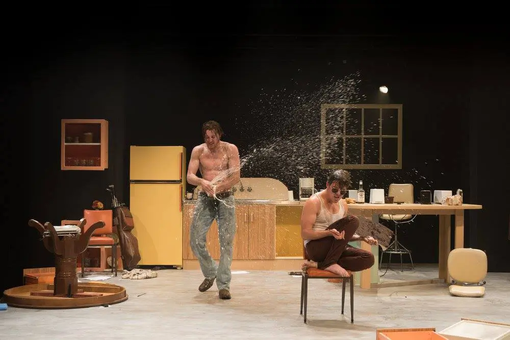 A theatre set: one man sprays champagne on another inside a partially destroyed kitchen