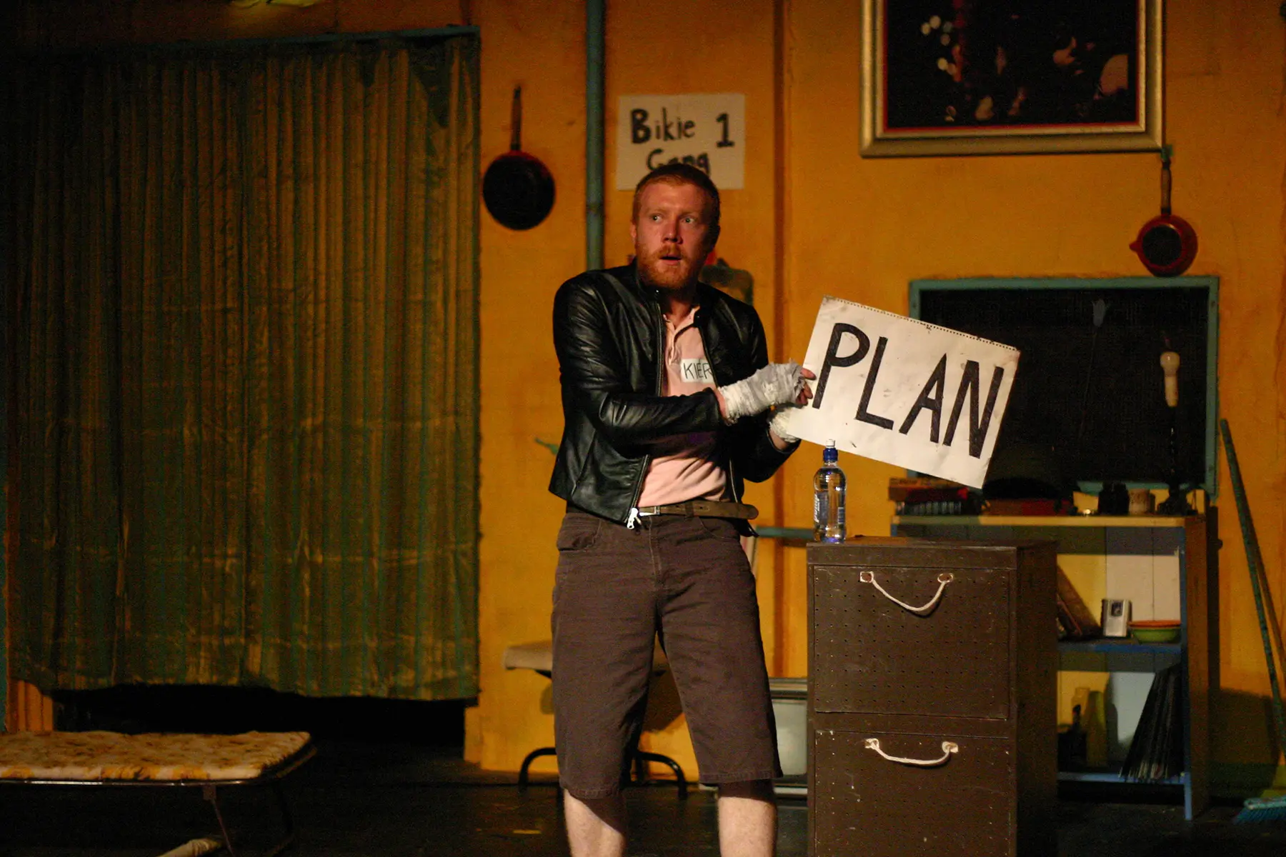 A man in a leather jacket holds a sign that says PLAN